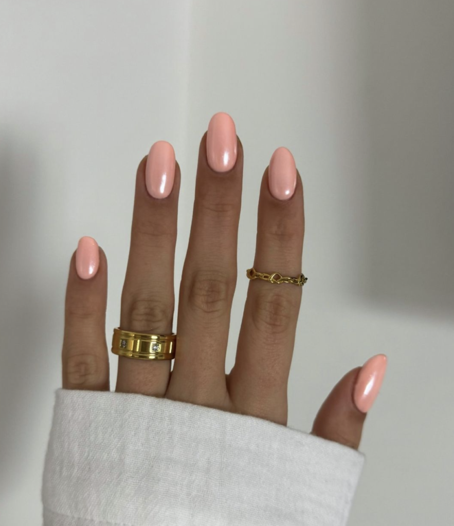 A hand adorned with almond-shaped nails painted in a glossy, warm peach shade, paired with a sophisticated gold ring.